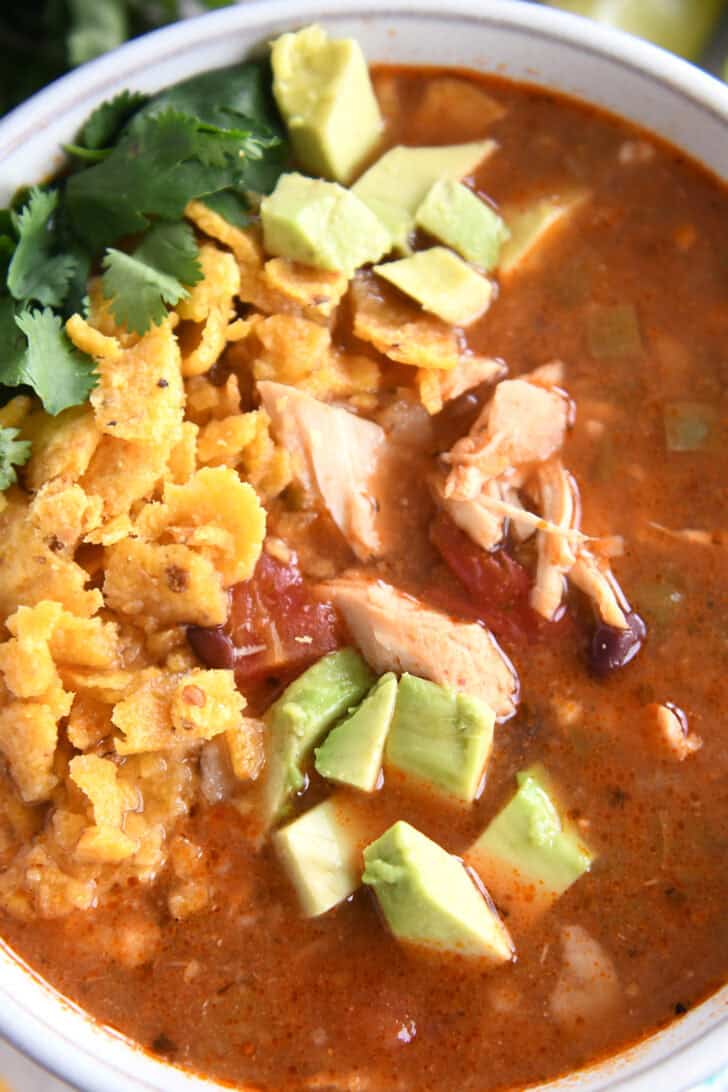 Top down view of a white bowl full of chicken tortilla soup topped with diced avocados, crushed chips, and chopped cilantro.