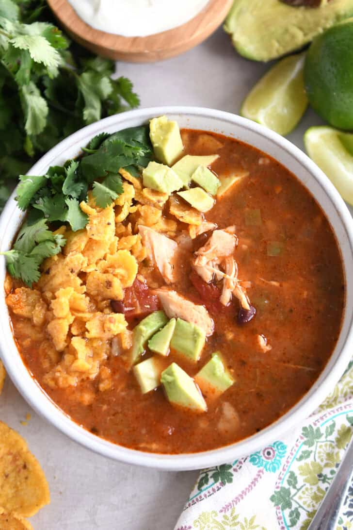 Top down view of a white bowl full of chicken tortilla soup topped with diced avocados, crushed chips, and chopped cilantro.