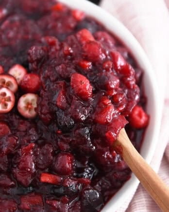 Wooden spoon scooping bite of cranberry chutney in white bowl.