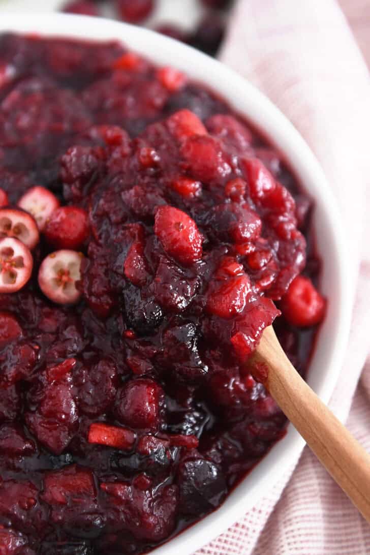 Wooden spoon scooping bite of cranberry chutney in white bowl.