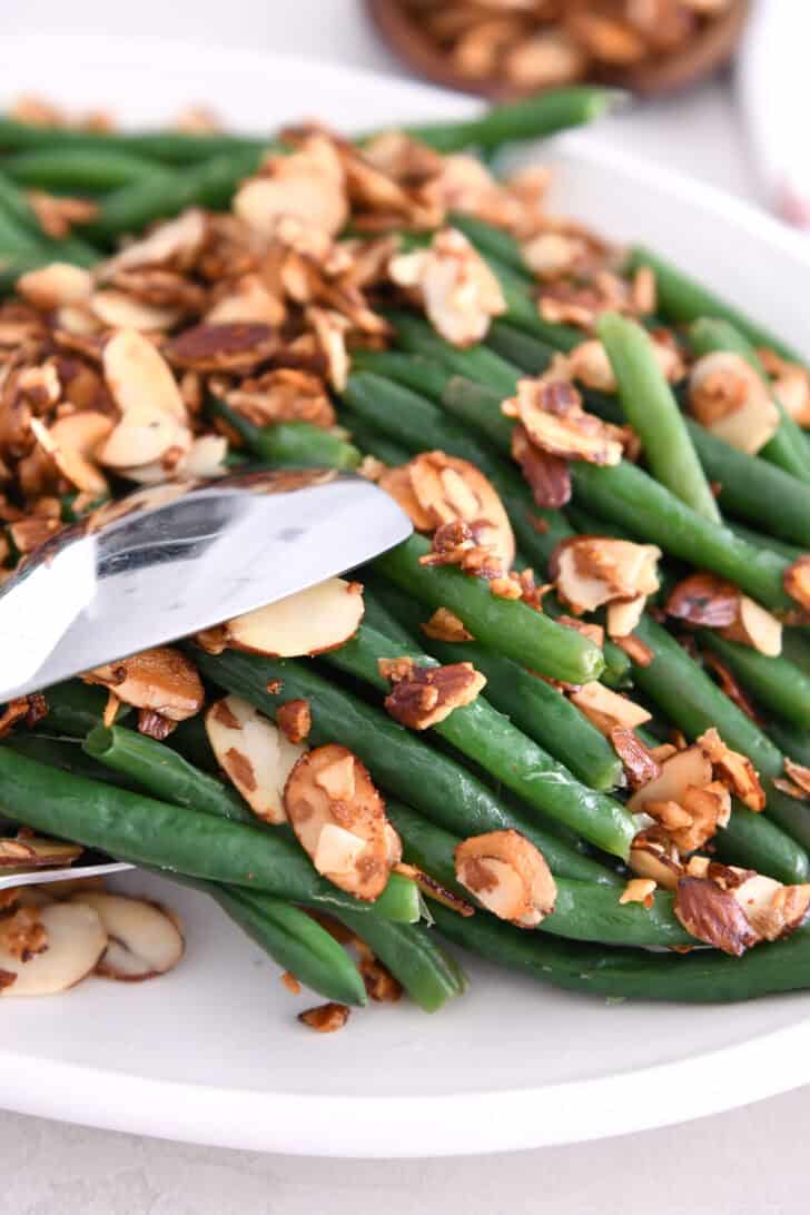 Tongs grabbing serving of steamed green beans with toasted almonds and garlic.
