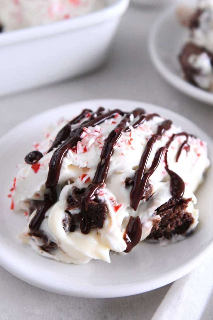 Serving of brownie peppermint bark pudding trifle on white plate drizzled with hot fudge sauce.