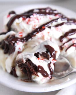 Spoon scooping bite of brownie pudding trifle on white plate drizzled with hot fudge sauce.