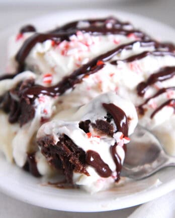 Spoon scooping bite of brownie pudding trifle on white plate drizzled with hot fudge sauce.