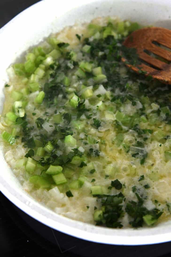 Celery, parsley, and onions cooking in butter in white skillet.