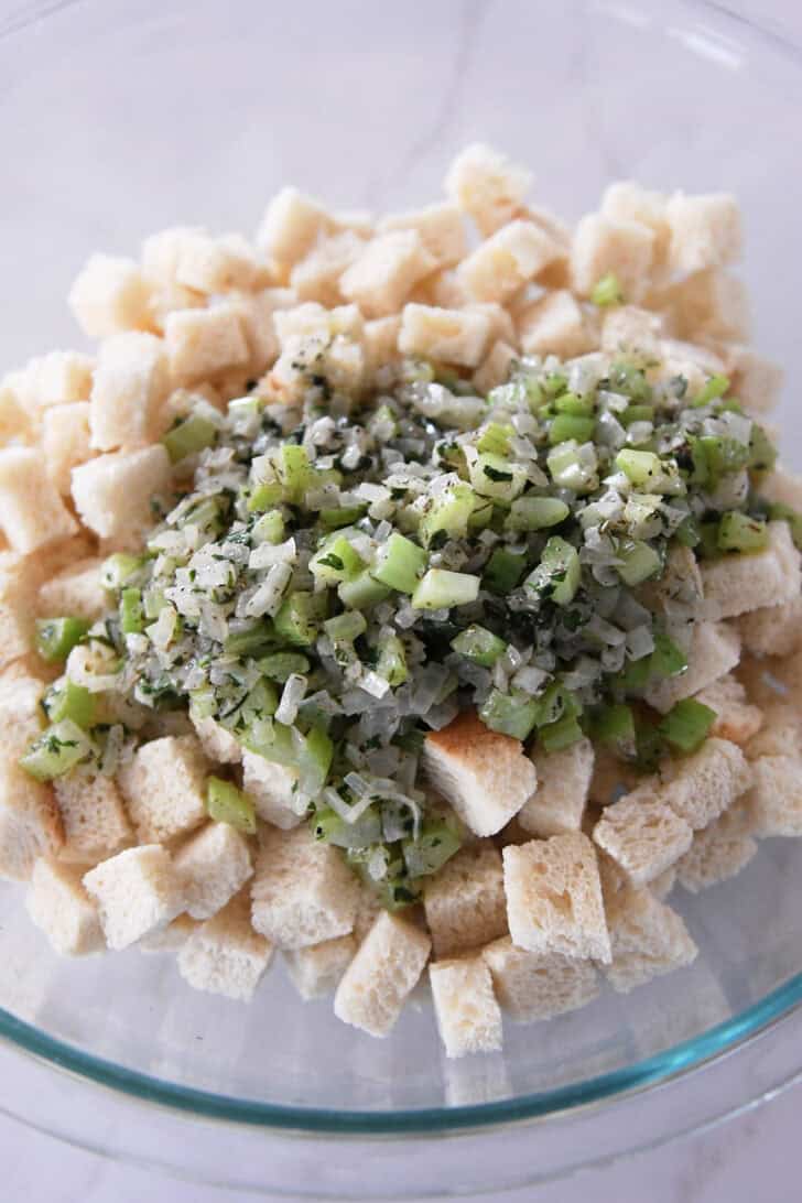 Sauteed onions and celery on top of toasted bread cubes in glass bowl.