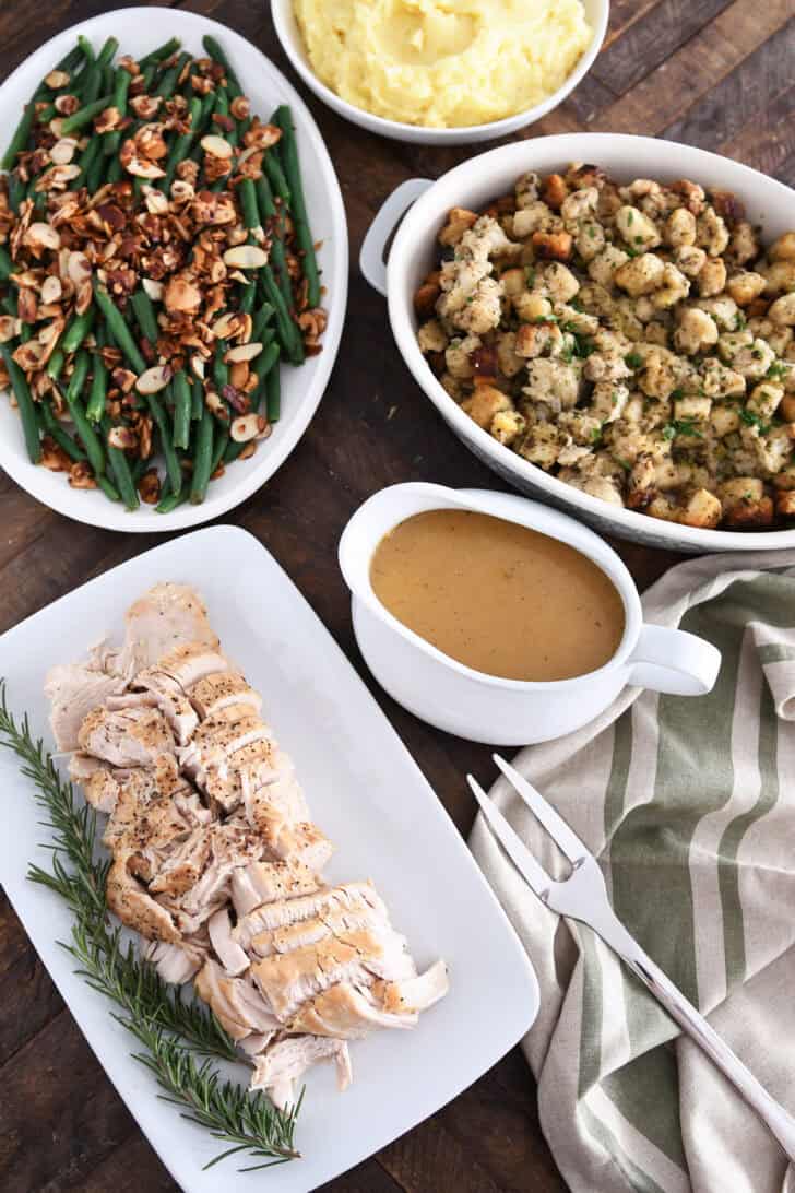 Top down view of platters of slow cooker turkey breast, gravy, stuffing, green beans and mashed potatoes.