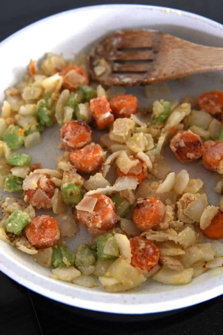 White skillet with wooden spoon and flour-coated carrots, celery and onions.