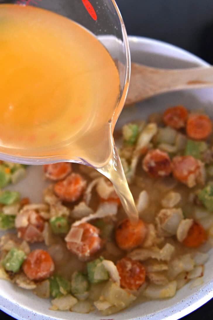 Pouring broth into white skillet with carrots, celery and onions.
