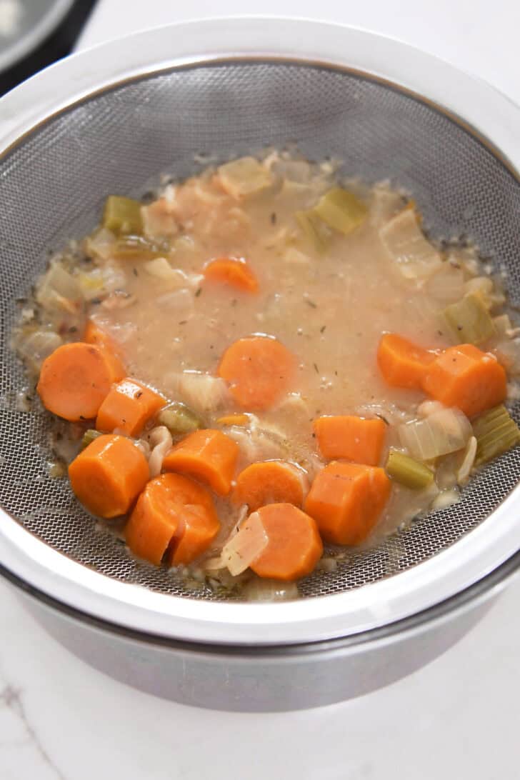 Fine mesh strainer over saucepan with cooked carrots, celery and broth mixture.