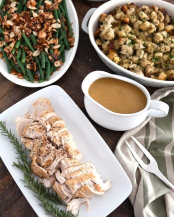 Top down view of platters of turkey, rosemary, gravy, stuffing, green beans and mashed potatoes.