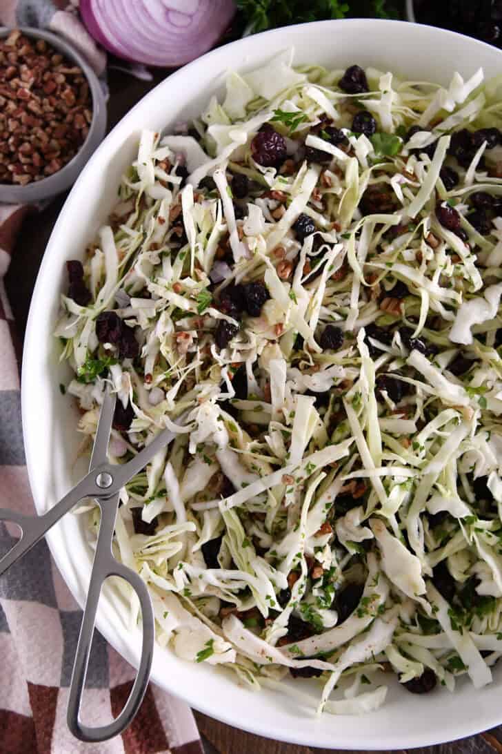 White bowl with salad tongs scooping portion of thin sliced green cabbage, dried cranberries, pecans and red onions.