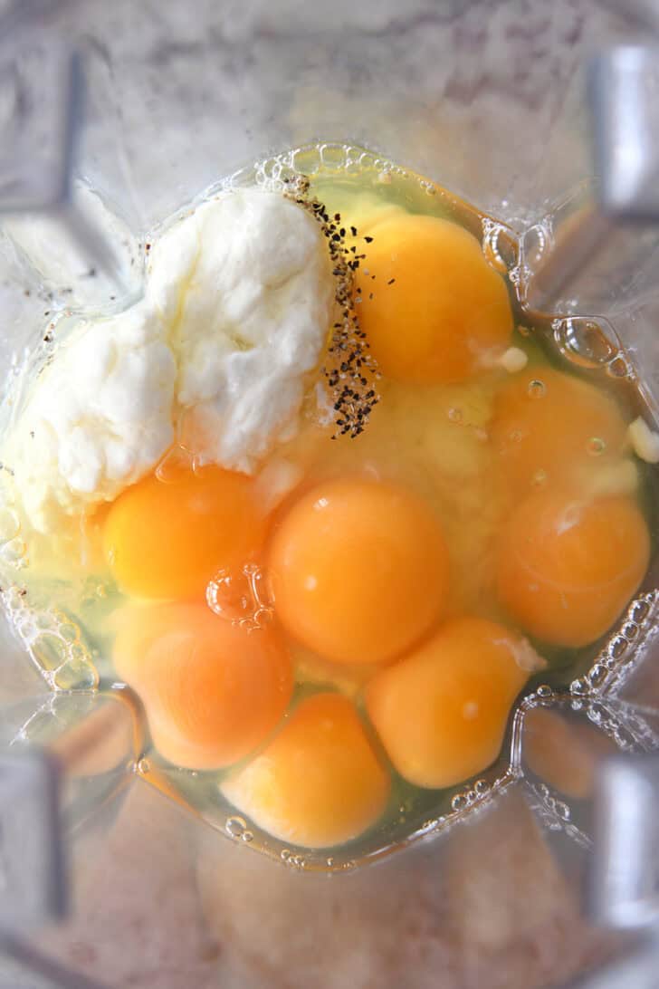 Top down view of a blender full of raw eggs, cracked black pepper, and cottage cheese.