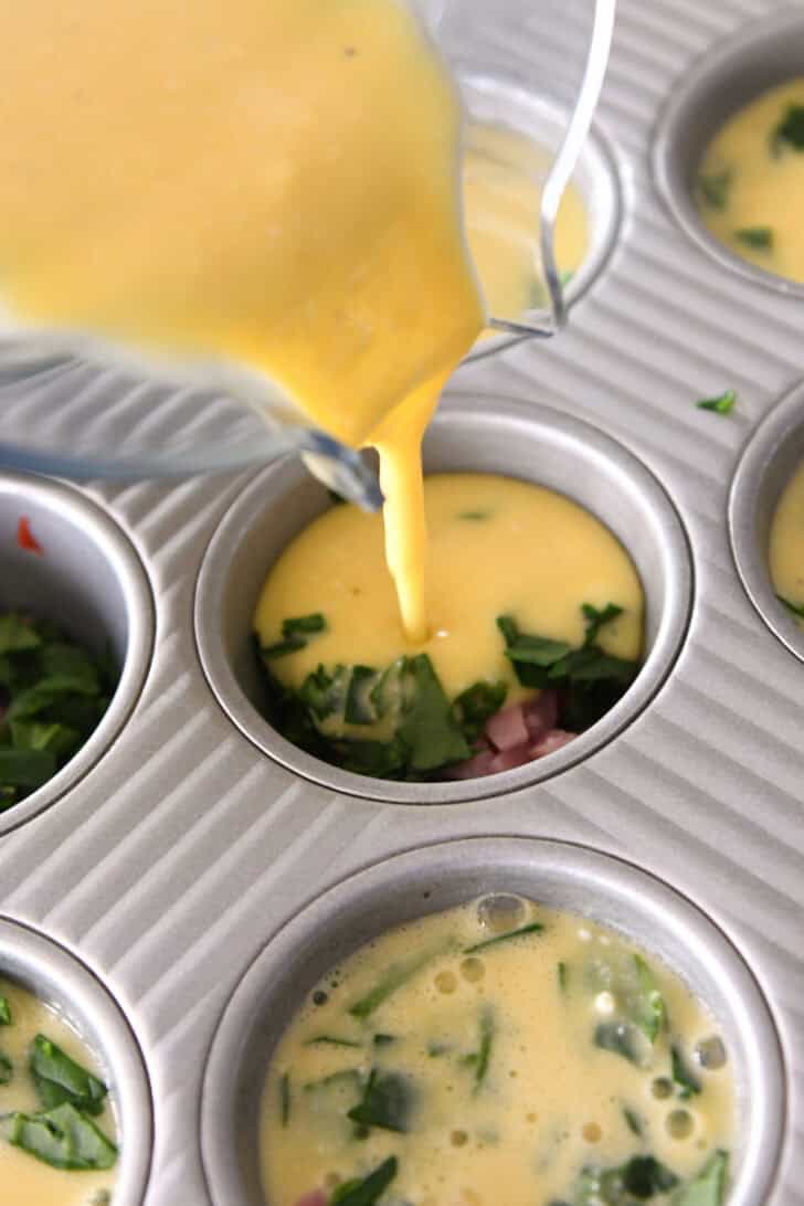 A glass measuring cup pouring raw egg into a muffin tin filled with chopped vegetables and diced ham.
