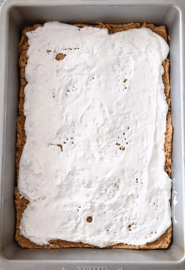 Top down view of a metal pan with a graham cracker crust base and a layer of gooey marshmallow spread on top.