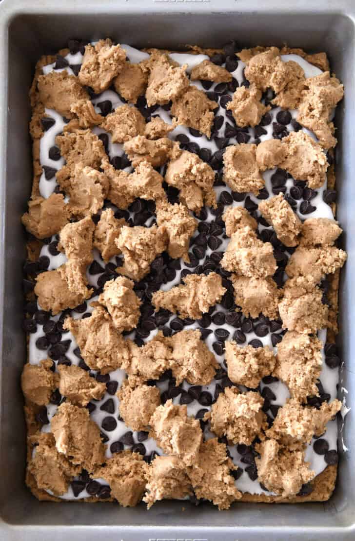 Top down view of a metal pan with dollops of graham cracker batter on top of chocolate chips and a layer of marshmallow.