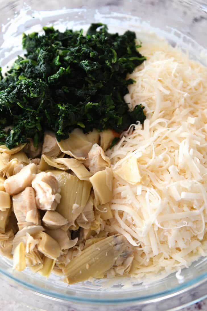 Top down view of a glass bowl with chopped artichokes, shredded cheese, and chopped spinach.