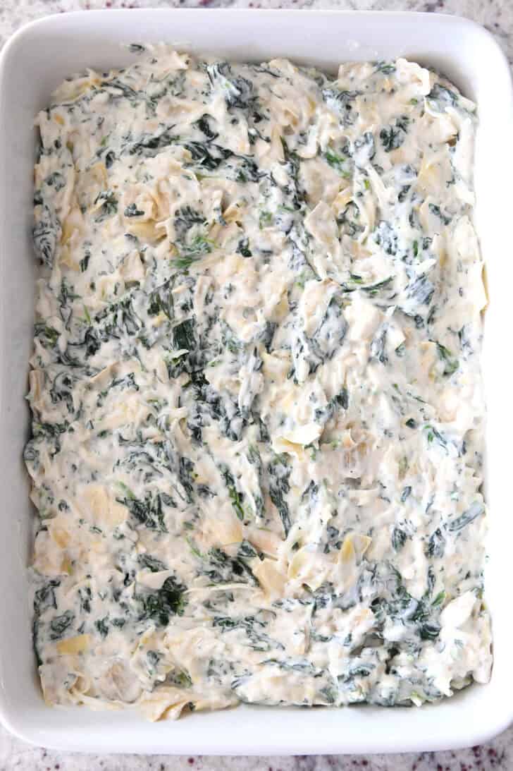 Top down view of a white casserole dish with uncooked creamy spinach artichoke dip.