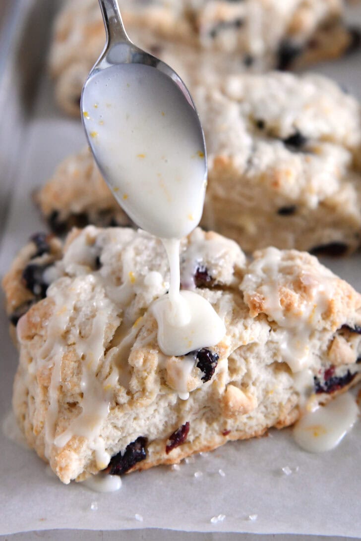 Spoon drizzling orange creamy glaze over baked scone.