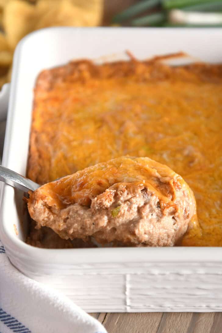 Serving spoon scooping serving of warm bean dip topped with cheese in white square baking dish.