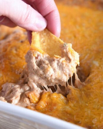 Fingers grabbing tortilla chip and dipping in cheesy, warm bean dip.