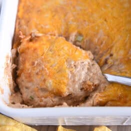 Serving spoon scooping serving of warm bean dip topped with cheese in white square baking dish.