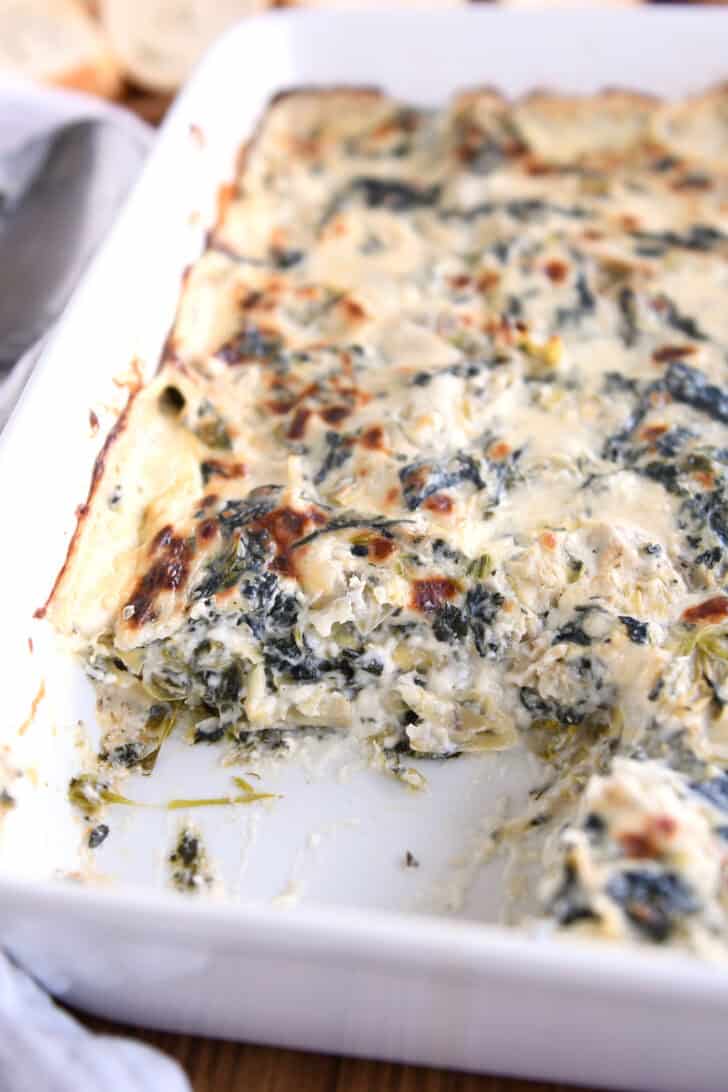A white casserole dish with a creamy cooked spinach artichoke dip. A scoop it taken out of the corner of the dish.