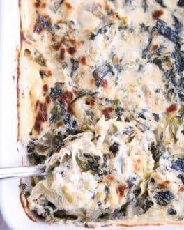 Top down view of a spoon taking a large scoop of spinach artichoke dip out of a white casserole dish full of dip.