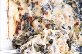 Top down view of a spoon taking a large scoop of spinach artichoke dip out of a white casserole dish full of dip.