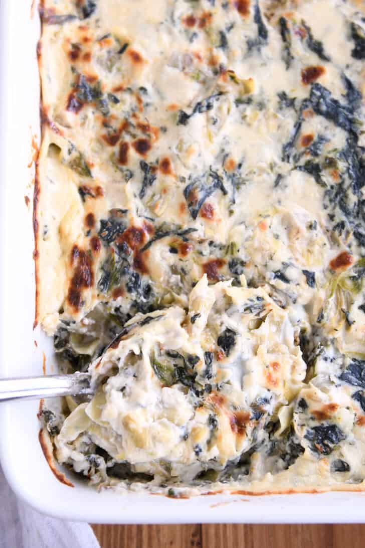 Top down view of a spoon taking a large scoop of spinach artichoke dip out of a white casserole dish full of dip.