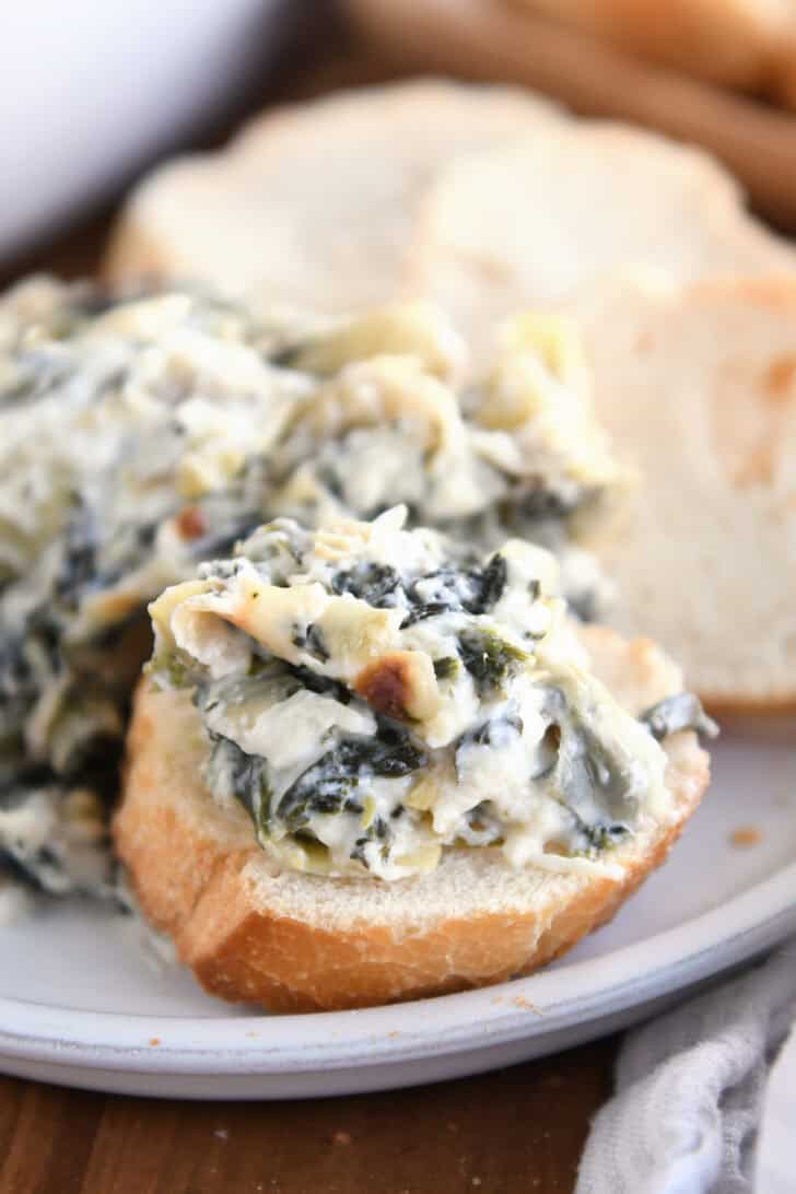 A small piece of sliced baguette with creamy spinach artichoke dip on top. More sliced bread and dip are in the background.