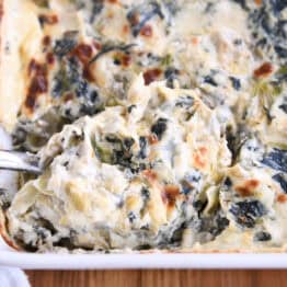 A large spoonful of creamy spinach artichoke dip being taken out of a white casserole dish full of the dip.