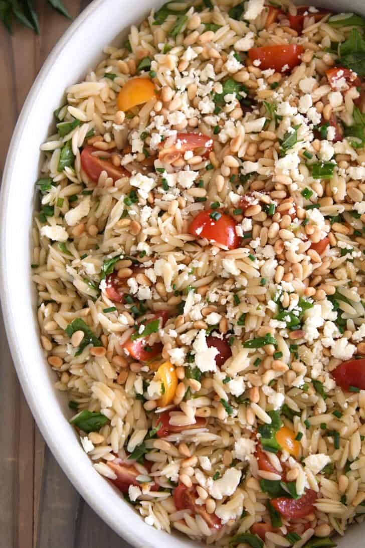Top down view of a white casserole dish with a cooked orzo salad with tomatoes, chopped basil, feta, and pine nuts.