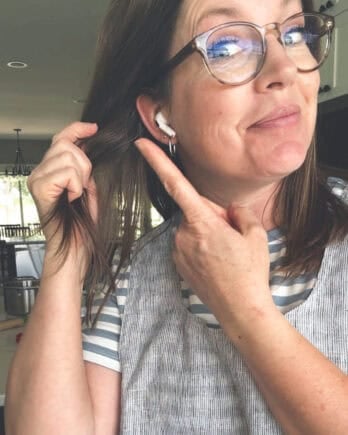 Woman in kitchen wearing apron pointing to earbuds in ear.