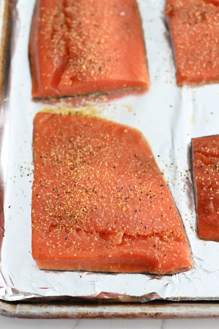 Salmon filets sprinkled with lemon pepper seasoning on foil-lined baking sheet.