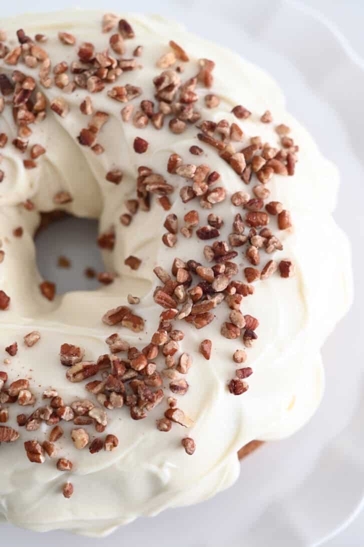 A carrot cake bundt cake covered in white frosting and sprinkled with chopped pecans on a white place.
