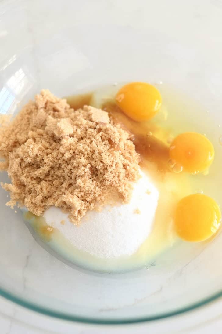 Top down view of a glass bowls with brown sugar, granulated sugar, and raw eggs.