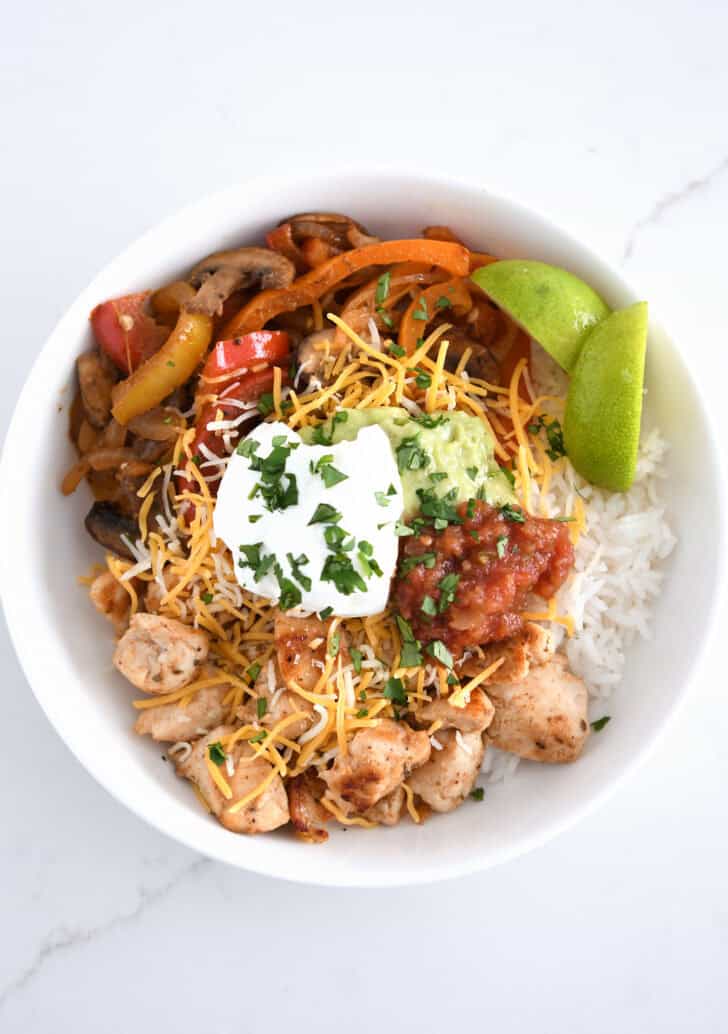 Top down view of rice, limes, grilled chicken, salsa, sour cream, guacamole, bell peppers and mushrooms.