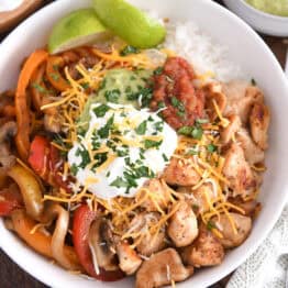 Top down view of white bowl with grilled chicken, peppers and onions, rice and limes.