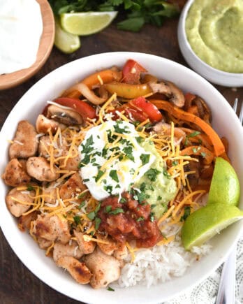 Top down view of rice, limes, grilled chicken, salsa, sour cream, guacamole, bell peppers and mushrooms.