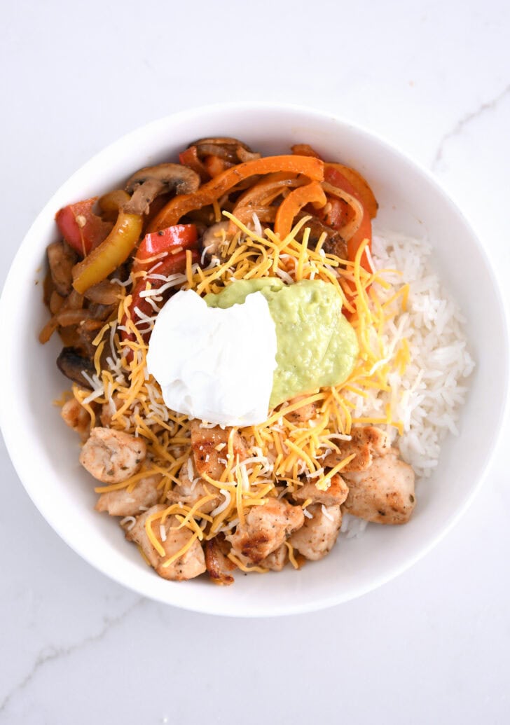 White shallow bowl with rice, cheese, grilled chicken, vegetables, sour cream and guacamole.
