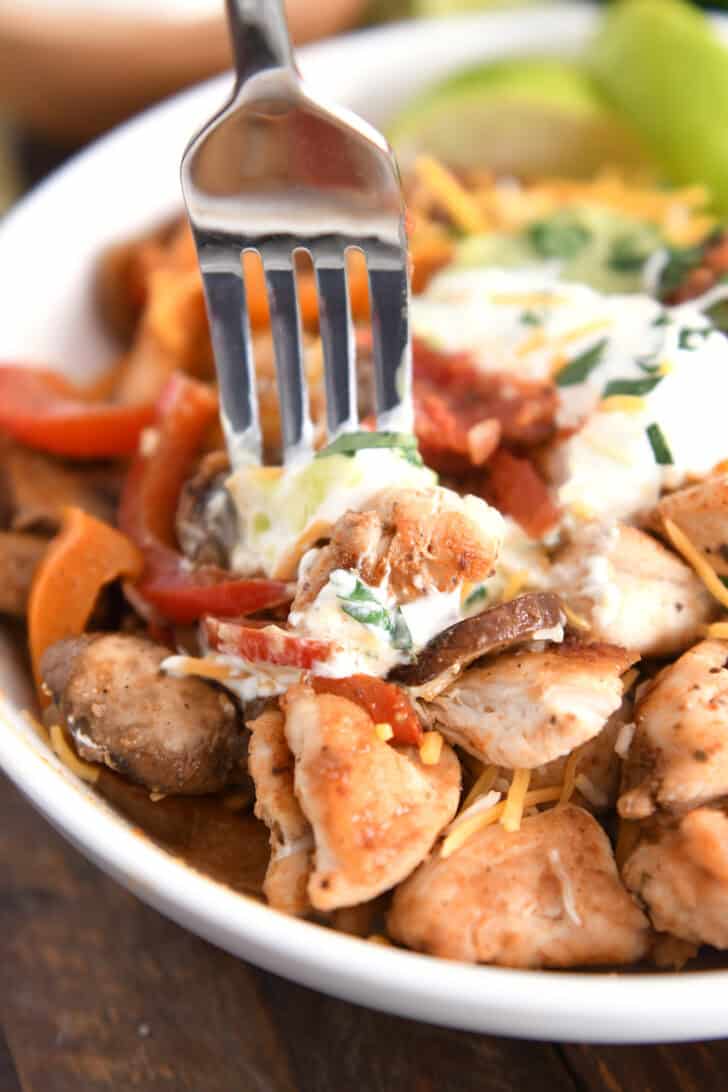 Fork scooping bite of grilled chicken, sautéed veggies, sour cream and guacamole.