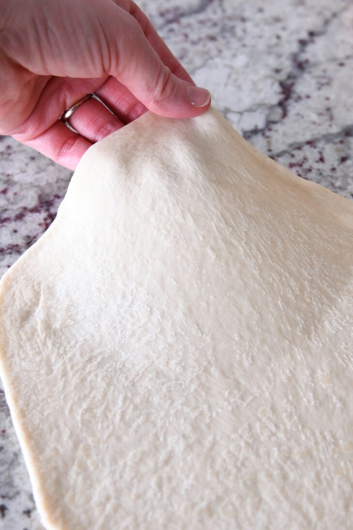 A hand pulling the side of a rolled out piece of bread dough up off the counter.