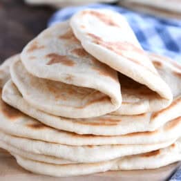 A stack of golden brown flatbread on a wooden cutting board. The top two flatbreads are folded in half. A blue and white checkered towel is laying beside the bread.