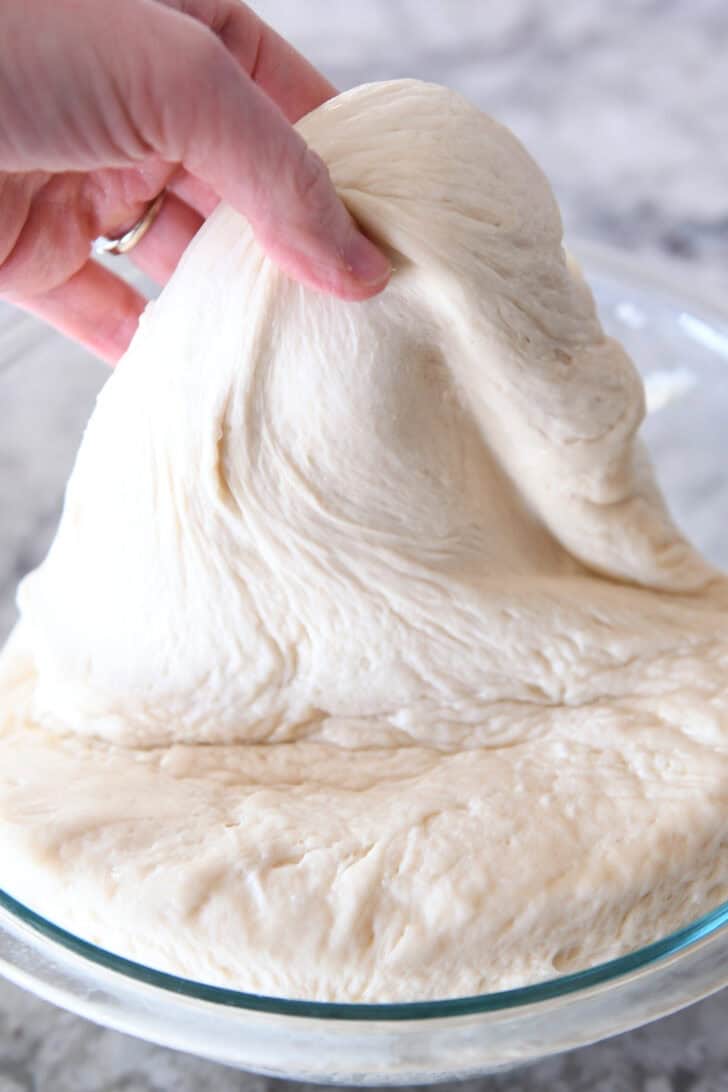 A hand pulling bread dough out of a glass bowl.
