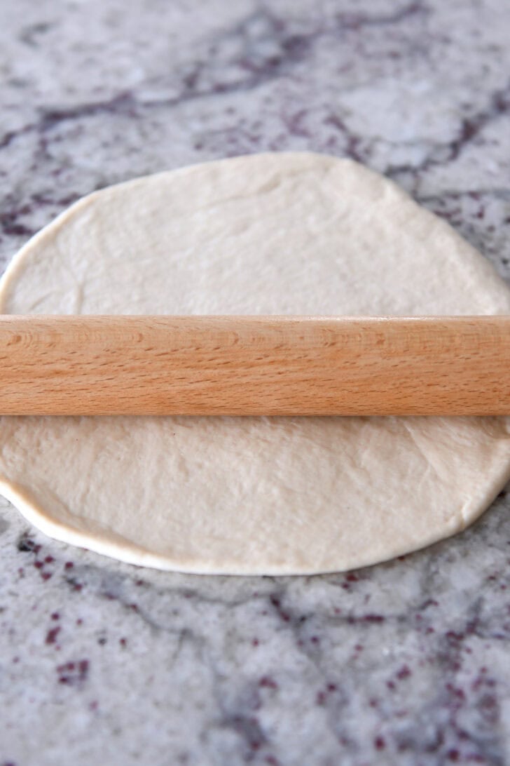 A wooden rolling pic rolling out bread dough into a circle on a granite countertop.