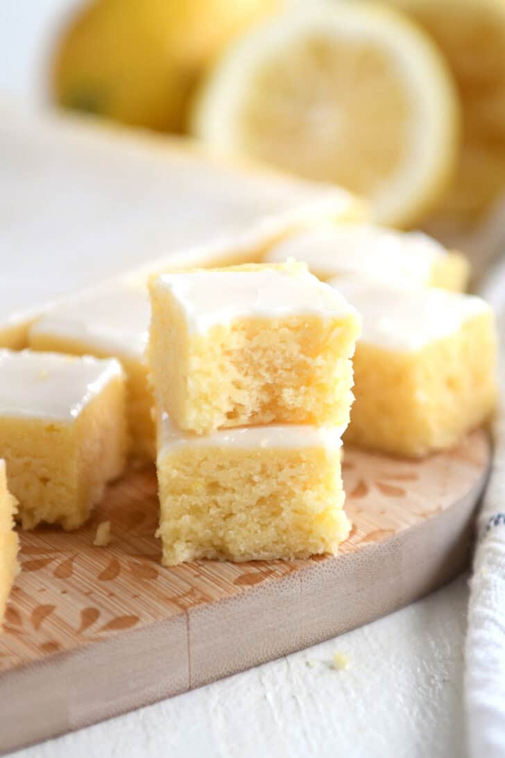 Two small lemon brownie squares with a white glaze topping stacked on top of each other on a wooden cutting board. More lemon brownie squares are in the background.