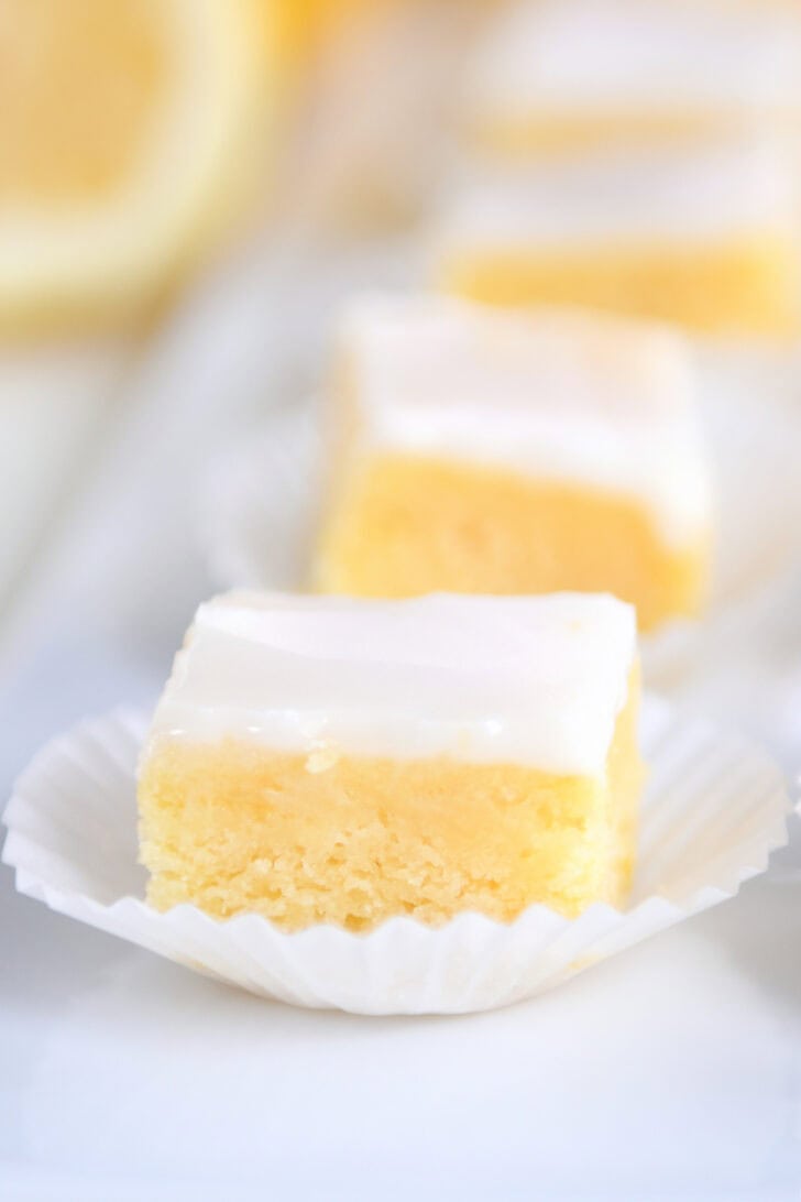 A line of lemon brownie squares topped with a white glaze in white muffin cups on a white serving platter.