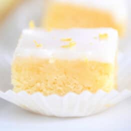A lemon brownie square topped with a white glaze and lemon zest sitting in a white muffin liner.