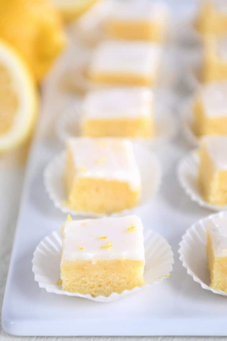 A line of lemon brownie squares topped with lemon zest and a white glaze in white muffin cups on a white serving platter.